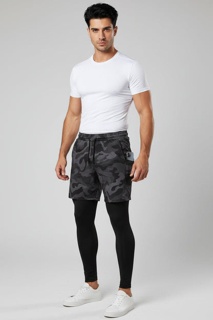 Man wearing a white t-shirt, black camouflage shorts, and black leggings on a light gray background