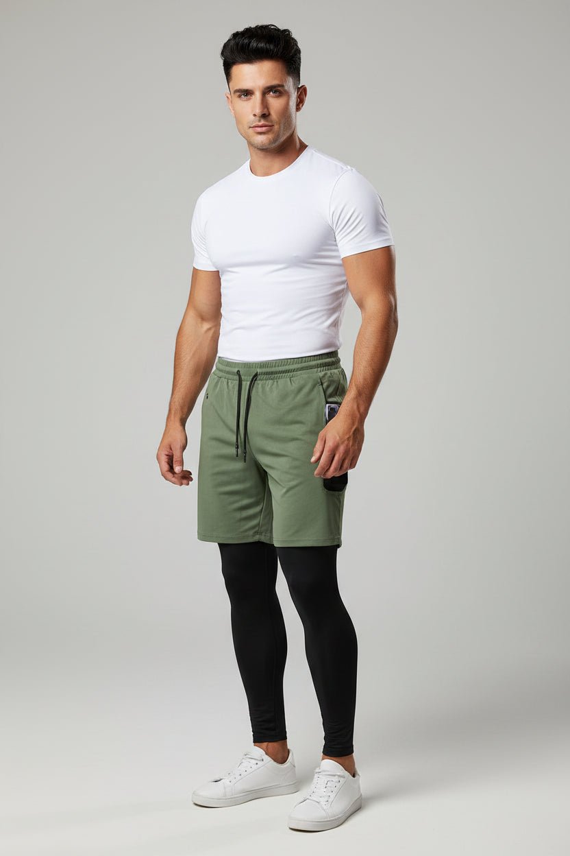 Man wearing a white t-shirt, green shorts, black leggings, and white sneakers on a gray background